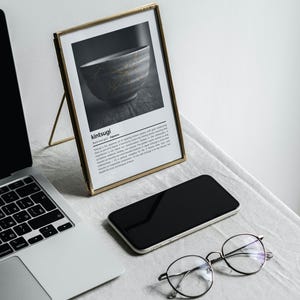 K&ouml;nnte beinhalten: Ein gerahmter Druck mit dem Wort "kintsugi" und einem Schwarz-Wei&szlig;-Bild einer Sch&uuml;ssel mit goldenen Akzenten. Ein Laptop, ein Smartphone und eine runde Brille befinden sich ebenfalls auf dem Tisch.