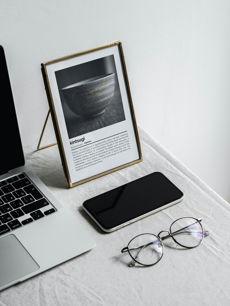 K&ouml;nnte beinhalten: Ein gerahmter Druck mit dem Wort "kintsugi" und einem Schwarz-Wei&szlig;-Bild einer Sch&uuml;ssel mit goldenen Akzenten. Ein Laptop, ein Smartphone und eine runde Brille befinden sich ebenfalls auf dem Tisch.