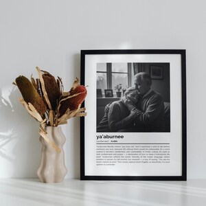 Op de afbeelding: Een zwart-witfoto in een lijst van een omhelzend stel, met het woord "ya'aburnee" onderaan. Links staat een beige vaas met gedroogde bloemen. De foto staat op een wit oppervlak.