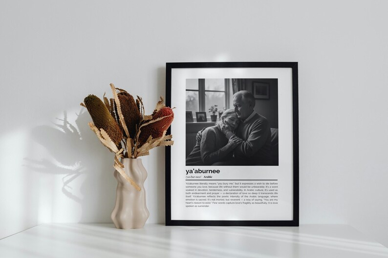 Op de afbeelding: Een zwart-witfoto in een lijst van een omhelzend stel, met het woord "ya'aburnee" onderaan. Links staat een beige vaas met gedroogde bloemen. De foto staat op een wit oppervlak.