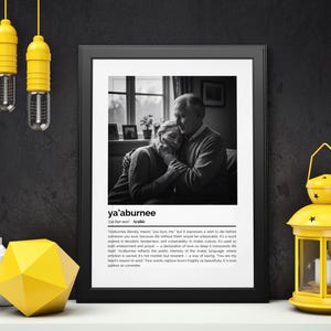 Op de afbeelding: Een zwart-wit afdruk met de tekst "ya'aburnee" en een afbeelding van een omhelzend stel. De afdruk wordt gepresenteerd met een gele lantaarn, kaarsen en een geel geometrisch object.