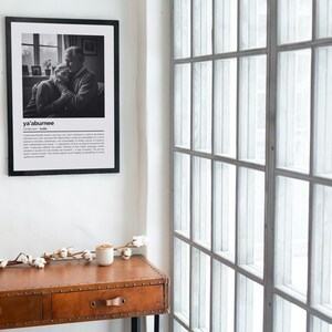 Op de afbeelding: Een zwart-wit print in een lijst aan een witte muur. De print toont een foto van twee mensen die elkaar omarmen, met het woord "ya'aburnee" boven een definitie. Een bruine houten tafel met lades en een witte kaars staan eronder.