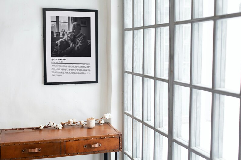 Op de afbeelding: Een zwart-wit print in een lijst aan een witte muur. De print toont een foto van twee mensen die elkaar omarmen, met het woord "ya'aburnee" boven een definitie. Een bruine houten tafel met lades en een witte kaars staan eronder.