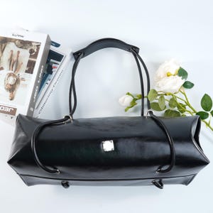 Może przedstawiać: Czarna skórzana torebka o prostokątnym kształcie i zaokrąglonych krawędziach. Torba ma dwa długie uchwyty i mały srebrny kwadratowy emblemat z przodu. Białe kwiaty i książki w tle.