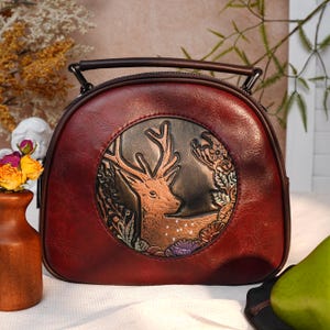 May include: A burgundy leather handbag with a circular embossed design featuring a deer. The bag has a top handle and a zipper closure. The deer is brown with white spots, surrounded by floral and leaf details.