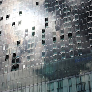 Puede incluir: Un edificio alto y moderno con una fachada de vidrio. El edificio refleja el sol y el cielo, creando un patrón abstracto brillante. El edificio tiene un gran letrero que dice "Hilton" en el lateral.