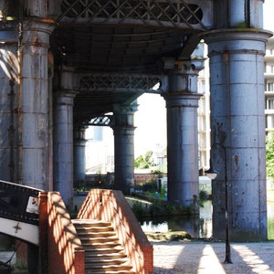 May include: A brick staircase leads up to a bridge under a large, gray, metal structure with pillars. The bridge is over a canal with water and green plants.