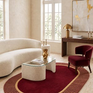 Puede incluir: Una alfombra ovalada con un centro rojo intenso y un borde ancho y texturizado con rayas verticales alternas en rojo y beige. La alfombra se coloca sobre un suelo claro, sirviendo de base para un sofá curvo de color crema y una silla roja.