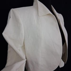 May include: A cream-colored blazer with a classic collar and tailored fit. The jacket is displayed on a black mannequin, showcasing its structured design and clean lines. The fabric appears to be a lightweight material.