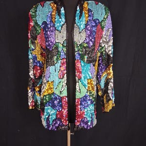 May include: A colorful sequined jacket with an open front. The jacket is covered in a variety of sequins in shades of blue, purple, red, green, gold, and black. The jacket is displayed on a mannequin against a black background.