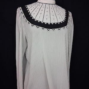 May include: A long-sleeved blouse with vertical black and white stripes. The blouse features a high collar and a decorative yoke with black and white geometric patterns. The yoke has a black border with white triangle accents and vertical lines.