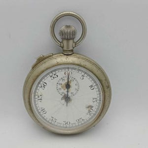Pode incluir: Um cronômetro prateado vintage com mostrador branco e algarismos pretos. O relógio apresenta um anel na parte superior e um mostrador com marcações de segundos e minutos. O mostrador do relógio apresenta algum desgaste.