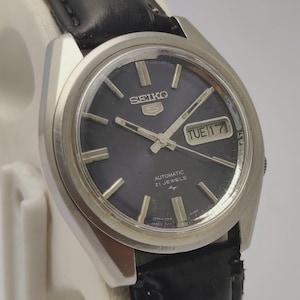 May include: A vintage Seiko 5 automatic wristwatch with a black dial, silver hands, and a day-date display. The watch has a stainless steel case and a black leather strap. The dial reads "Automatic 21 Jewels".