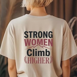 May include: A beige t-shirt with the text "STRONG WOMEN CLIMB HIGHER" in black, pink, and burgundy. The design includes a mountain range graphic and decorative elements. The person is facing away from the camera.