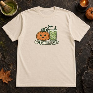 May include: A cream-colored short-sleeve t-shirt featuring a graphic of an orange jack-o'-lantern pumpkin next to a glass of green liquid with ice cubes and a straw. A small black bat is above the glass, and the text OCTOBER SIPS is below.