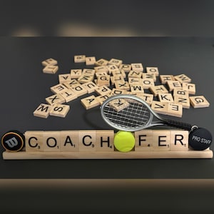 May include: A tennis-themed decorative display featuring wooden letter tiles spelling "COACH FER", a miniature tennis racket, a tennis ball, and a Wilson logo. A black Pro Staff keychain and scattered letter tiles complete the arrangement.