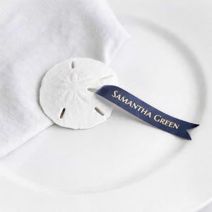 May include: A white sand dollar place card holder with a navy blue ribbon. The ribbon has the name "SAMANTHA GREEN" printed in gold. The sand dollar is resting on a white plate next to a white linen napkin.