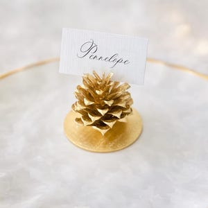 May include: A gold-painted pinecone place card holder with a circular base, featuring a white rectangular card with the name Penelope written in black cursive script, centered on a white plate with a gold-toned scalloped rim.