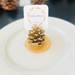 May include: A gold-painted pinecone place card holder with a white card. The card has the name "Mathew Reese" and "Table eight" printed on it, surrounded by a gold leaf design. The holder sits on a gold base and a white plate.
