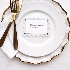 May include: A place setting with a white plate, gold-rimmed charger, and gold flatware. A white napkin and a name card with the text "Elizabeth Morris tabletwo" are on the plate. The setting is on a white surface.