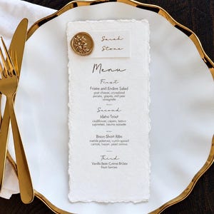 May include: A white menu card with deckled edges featuring black text listing courses including Frisée and Endive Salad, Idaho Trout, Bison Short Ribs, and Vanilla Bean Crème Brûlée, topped with a gold wax seal and a name card reading Sarah Stone.