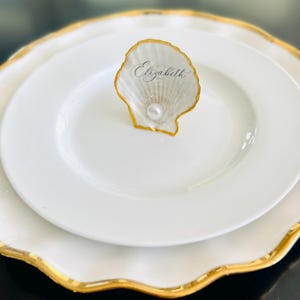 May include: A white plate with a gold rim holds a smaller white plate. A seashell place card holder with the name "Elizabeth" sits on top. The seashell is gold-edged and has a pearl accent.