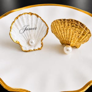 May include: Two seashell-shaped place card holders on a white plate with a gold rim. One shell is white with gold trim and the name "James" written on it. The other shell is gold. Each shell has a pearl.