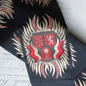 May include: Black fabric ribbon with gold and red embroidered crests. The crests feature a lion, a flower, and a shield with a pattern. The text "FOULARD" and "BASQUE" is embroidered on the ribbon.