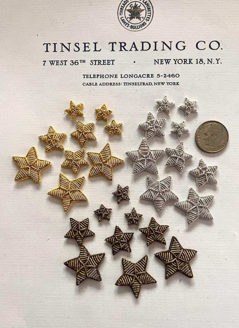 K&ouml;nnte beinhalten: Sortiment aus goldenen, silbernen und bronzenen sternf&ouml;rmigen Verzierungen. Die Sterne sind aus gewebtem Garn gefertigt und haben verschiedene Gr&ouml;&szlig;en. Der Text "TINSEL TRADING CO. 7 WEST 36TH STREET NEW YORK 18, N.Y. TELEPHONE LONGACRE 5-2460 CABLE ADDRESS: TINSELTRAD, NEW YORK" ist auf wei&szlig;em Hintergrund gedruckt.