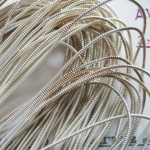 May include: A close-up of a spool of silver and gold metallic cord. The cord is thin and has a slightly textured surface.