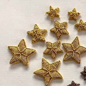 K&ouml;nnte beinhalten: Zehn goldene sternf&ouml;rmige Verzierungen, jede mit einem gewebten Design. Die Sterne sind in einem zuf&auml;lligen Muster auf einem wei&szlig;en Hintergrund angeordnet.