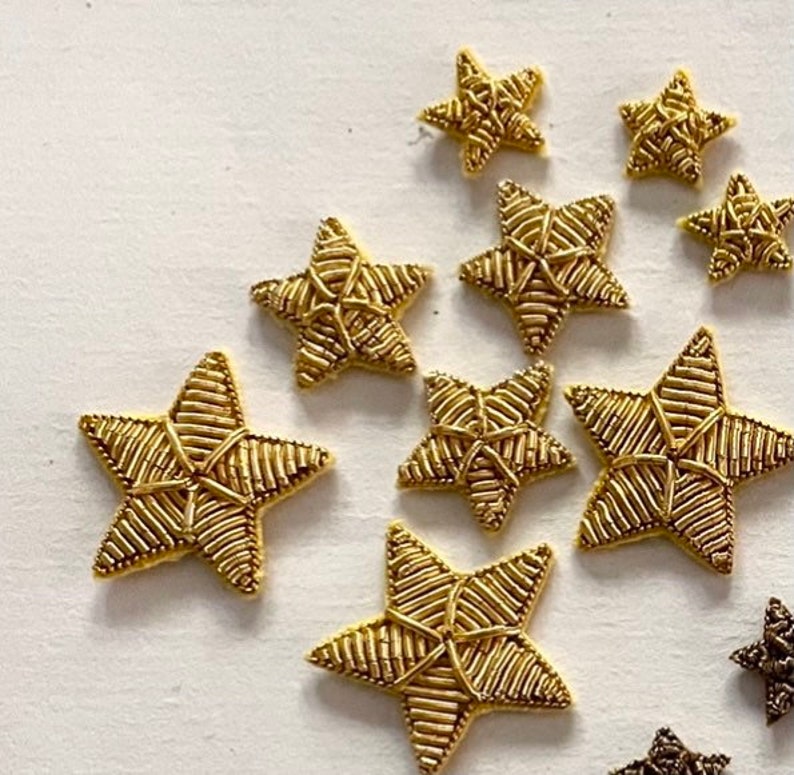 K&ouml;nnte beinhalten: Zehn goldene sternf&ouml;rmige Verzierungen, jede mit einem gewebten Design. Die Sterne sind in einem zuf&auml;lligen Muster auf einem wei&szlig;en Hintergrund angeordnet.