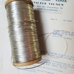 May include: A spool of silver wire wound around a wooden bobbin. The spool is labeled "Etablissements "VINOX"" and "VICTOR VIGNON".