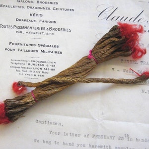May include: A bundle of gold metallic thread with red yarn at each end. The thread is tied together with a pink rubber band. The thread is on a white piece of paper with text that reads "Claude Pro" and "TINSEL TRADING CO."