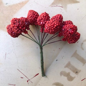 May include: A bundle of artificial berries in a vibrant red hue. The berries are clustered together, with a textured, bumpy surface. The stems are dark green and wrapped together with wire, creating a decorative floral accent.