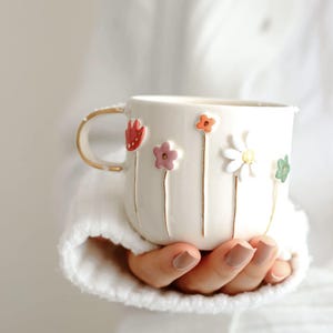 May include: Cream-coloured ceramic mug decorated with raised, colourful flower designs. The flowers are red, pink, orange, white, and green, with gold stems. The handle is gold. A decorative floral mug.