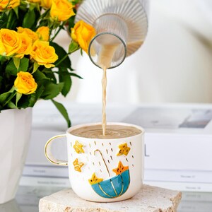 Puede incluir: Una taza de cerámica blanca con un asa dorada y un diseño de paraguas azul con estrellas amarillas se está llenando con un líquido de color claro. Rosas amarillas en un jarrón blanco en el fondo.