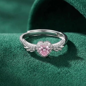 May include: A silver ring with a heart-shaped pink gemstone at the center. The ring features small, clear stones around the heart and on the wings that extend from the sides. The ring is set against a green background.