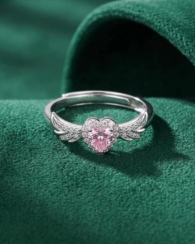 May include: A silver ring with a heart-shaped pink gemstone at the center. The ring features small, clear stones around the heart and on the wings that extend from the sides. The ring is set against a green background.