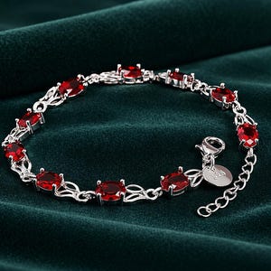 Könnte beinhalten: Ein silbernes Armband mit ovalen, tiefroten Edelsteinen. Das Armband hat ein filigranes Kettendesign, einen Karabinerverschluss und einen kleinen, runden Anhänger. Die Edelsteine sind in Silbermetall gefasst.