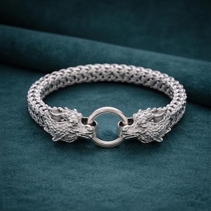 May include: A silver bracelet featuring two dragon heads facing each other, connected by a circular clasp. The bracelet has a woven chain design. The dragon heads have detailed scales and facial features. The bracelet is displayed on a dark teal surface.