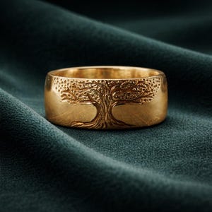 May include: A gold ring featuring a detailed tree design. The tree is etched into the band, showing the trunk, branches, and leaves. The ring is set against a dark green background, highlighting the intricate details of the jewellery.