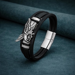 May include: Black leather bracelet with a silver eagle emblem. The bracelet has a magnetic clasp and two silver bands. The eagle emblem is detailed with feathers and a head.