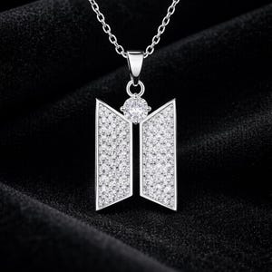 Pode incluir: Um colar com pingente prateado com um design geométrico. O pingente tem dois painéis verticais e um detalhe central em forma de diamante, todos cobertos por pequenas pedras brilhantes. O colar pende de uma corrente delicada sobre um fundo preto.