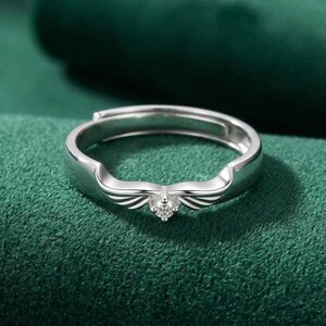 May include: A silver-colored ring with a wing design and a small, clear gemstone at the center. The ring is set against a dark green background, highlighting its metallic sheen and intricate details. The ring is adjustable.