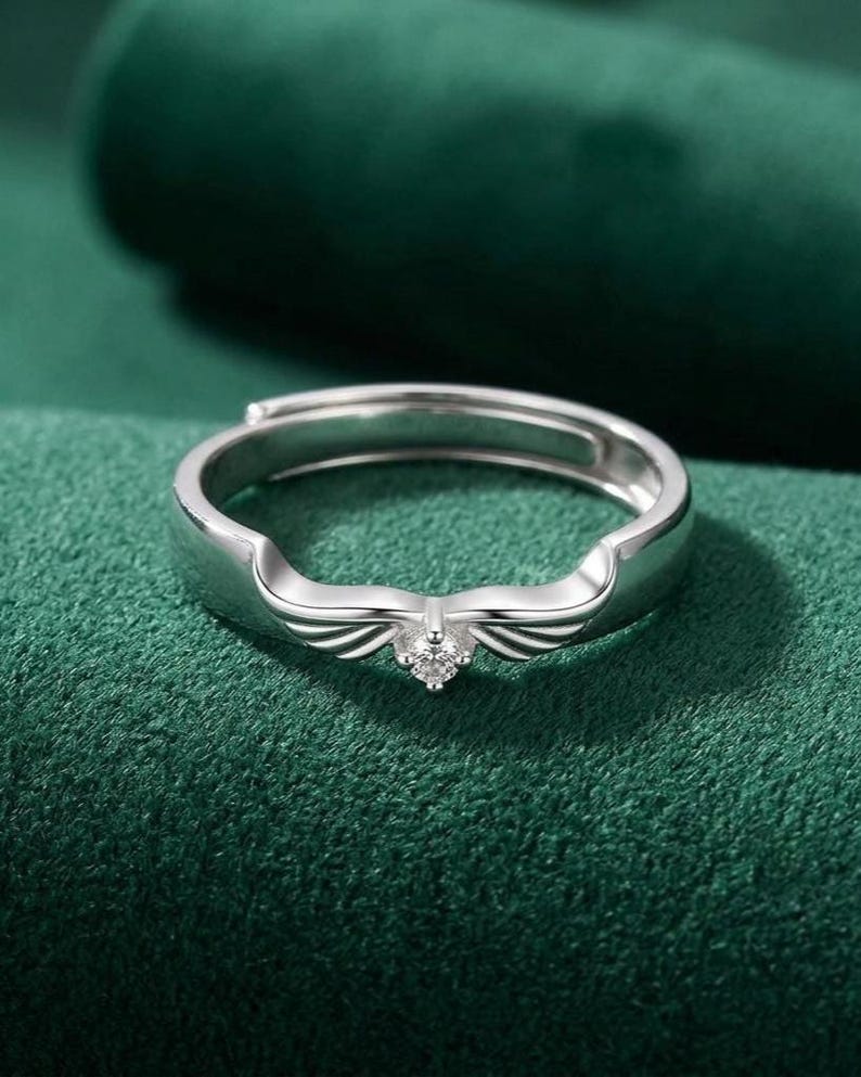 May include: A silver-colored ring with a wing design and a small, clear gemstone at the center. The ring is set against a dark green background, highlighting its metallic sheen and intricate details. The ring is adjustable.