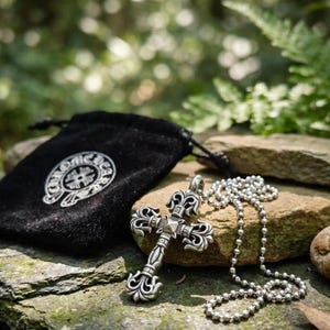 Peut inclure: Un pendentif croix en argent avec des volutes complexes et un clou pyramidal central, attaché à un collier à chaîne à billes en argent. Le pendentif est présenté à côté d'une pochette noire en velours avec un logo circulaire blanc CHROME HEARTS.