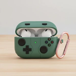 Könnte beinhalten: Grüne Schutzhülle für kabellose Ohrhörer, die einem Videospiel-Controller nachempfunden ist. Die Hülle hat ein kreuzförmiges Steuerkreuz, Tasten und die Wörter "SELECT" und "START". Ein roségoldener Karabinerhaken ist befestigt.