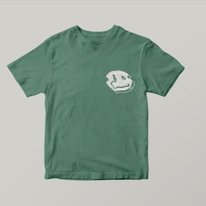 May include: A green t-shirt featuring a white, abstract smiley face design on the chest. The shirt is a solid, muted green colour and has short sleeves. The graphic is a distorted, outlined face.