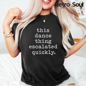 Puede incluir: Camiseta negra con el texto blanco "this dance thing escalated quickly". La camiseta tiene cuello redondo y mangas cortas. El logo de Retro Soul está en la esquina superior derecha.
